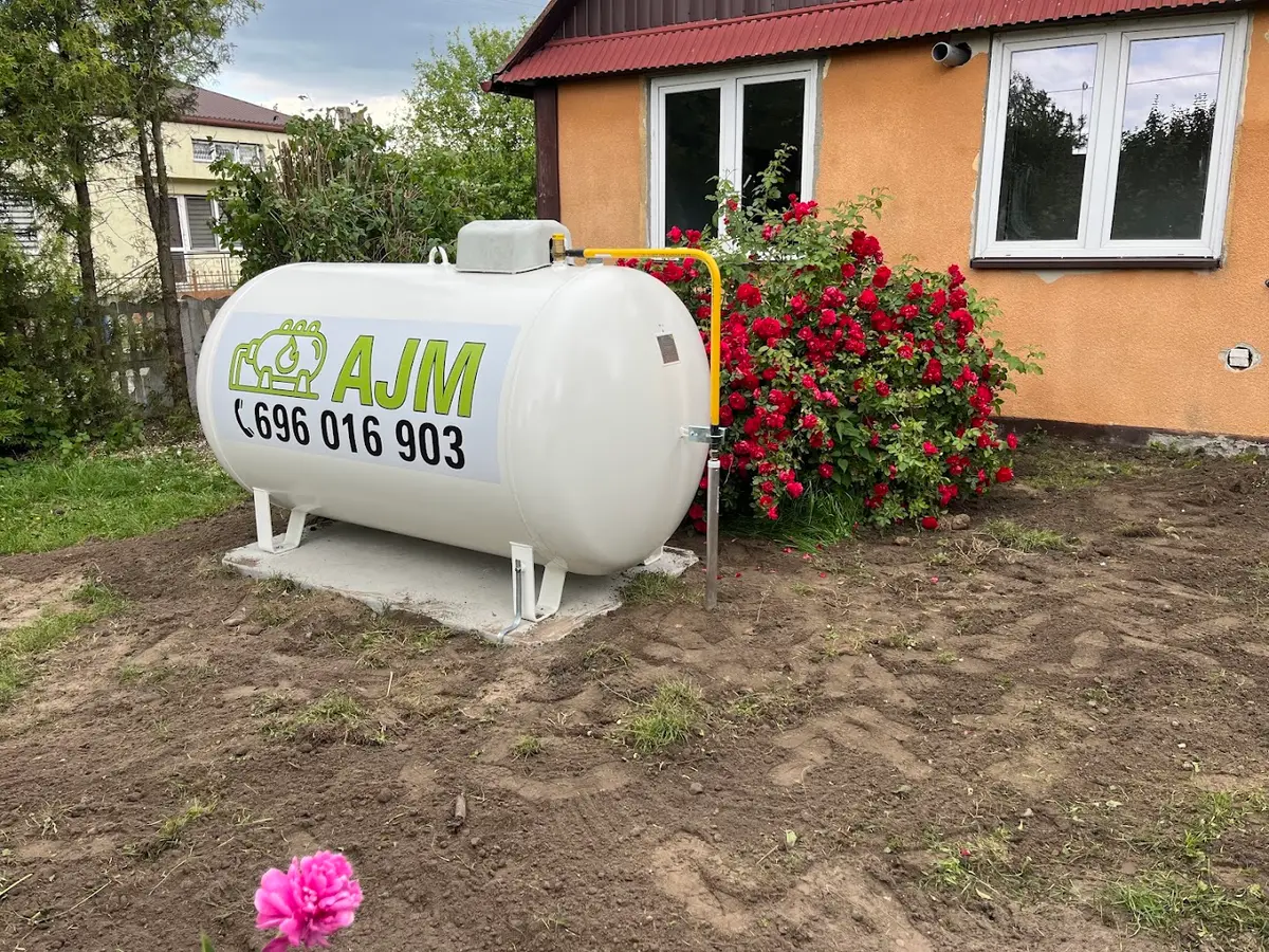 Biały zbiornik LPG z naklejką AJM przy domu jednorodzinnym, obok krzew czerwonych róż